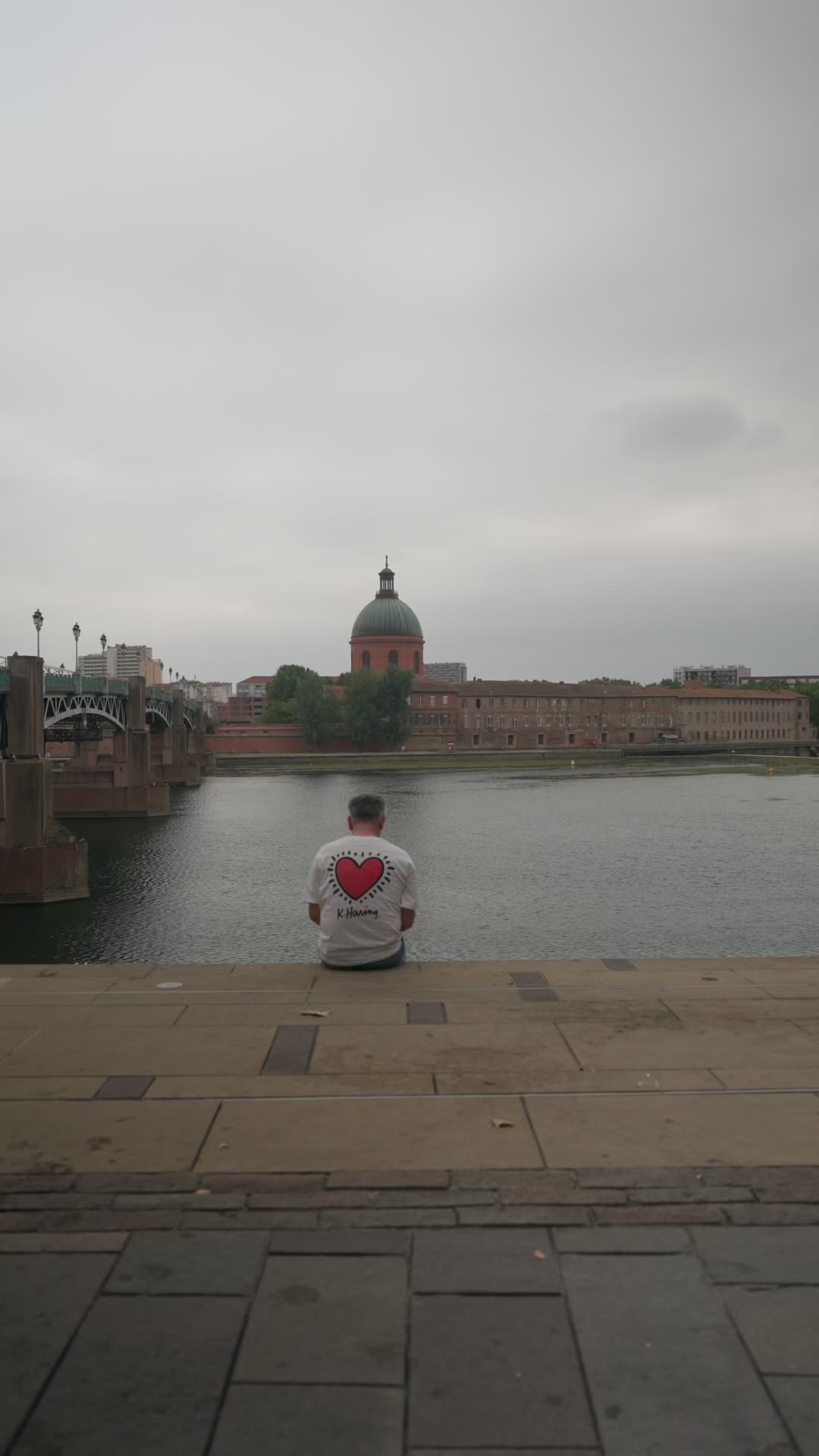 Melv de dos, assis sur le quai face à la Garonne et au Dôme de la Grave, un cœur rouge dessiné sur son tee-shirt